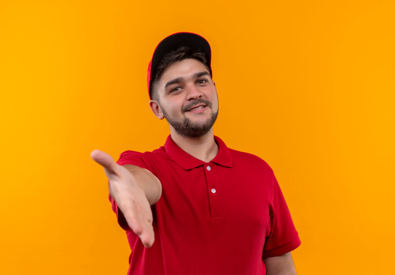young delivery man in red uniform and cap smiling friendly greeting offering hand standing over orange background